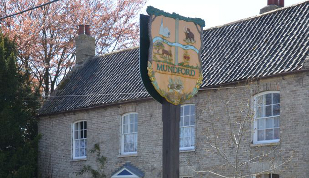 The Mundford village sign mounted on a tall post in front of a traditional brick cottage